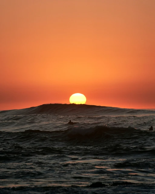 Ocean waves crash under an orange sunset with silhouettes of people in the water, creating a tranquil and scenic seascape.