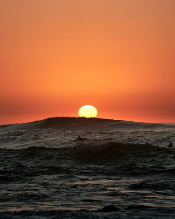Ocean waves crash under an orange sunset with silhouettes of people in the water, creating a tranquil and scenic seascape.