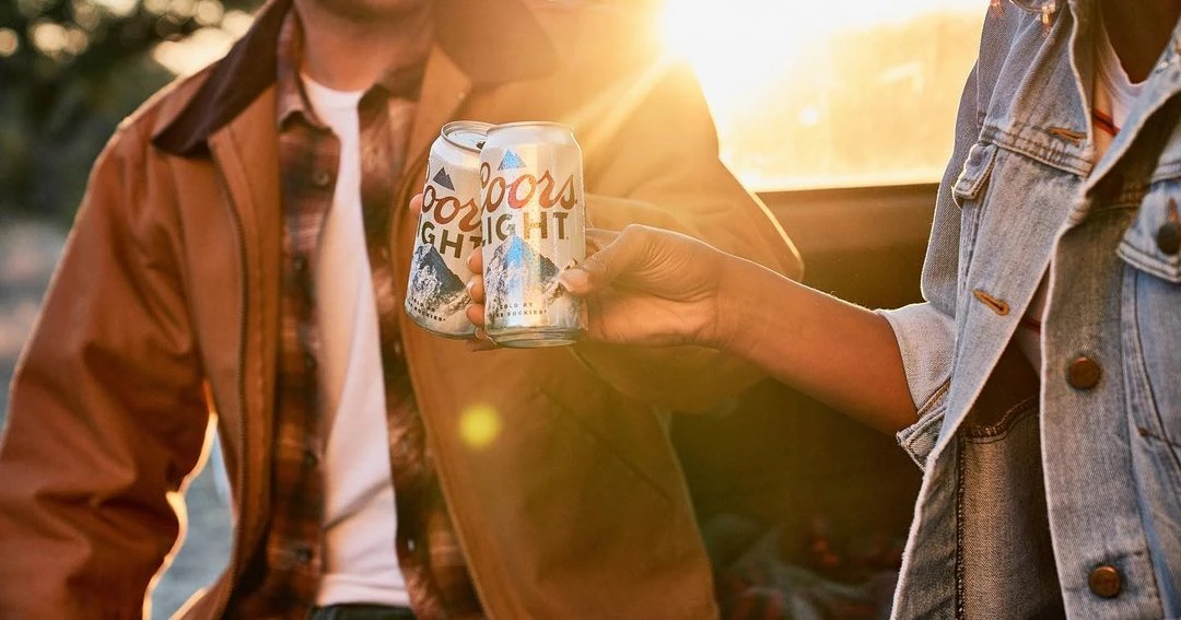 Coors Light beer cans are held by two adults with a denim jacket, and brown coat, and a bright sun is behind them.