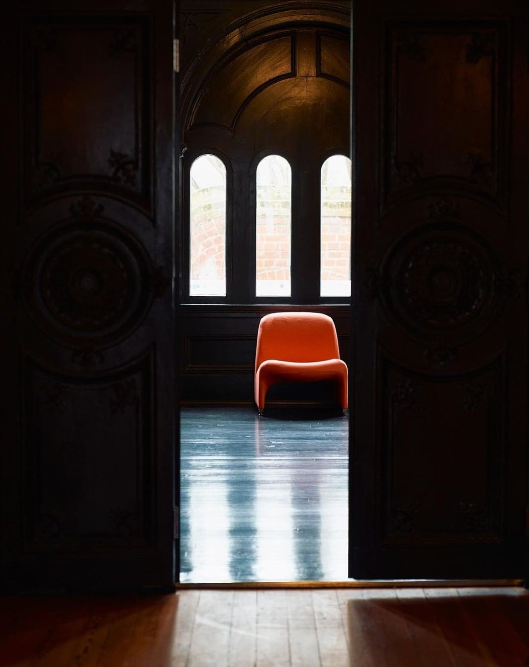 View through dark wooden doorway of orange armchair in a room with three arched windows, creating a compelling visual contrast.