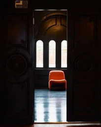 An orange armchair centered in a doorway looking into an arched room with dark wooden paneling and arched windows.