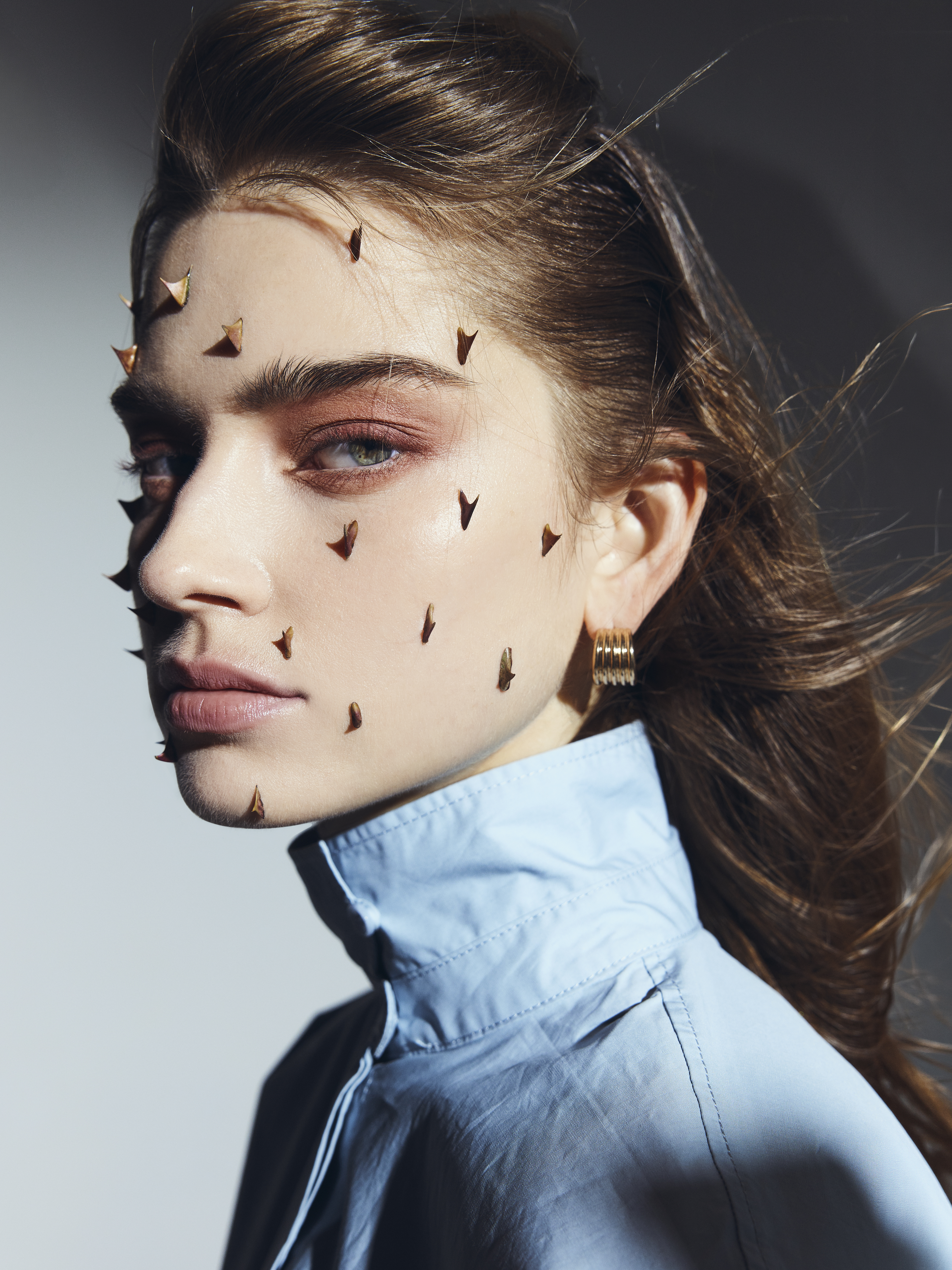 Close up portrait of a model with small decorative spikes on their face, wearing a light blue high-neck shirt, captured in Paris.