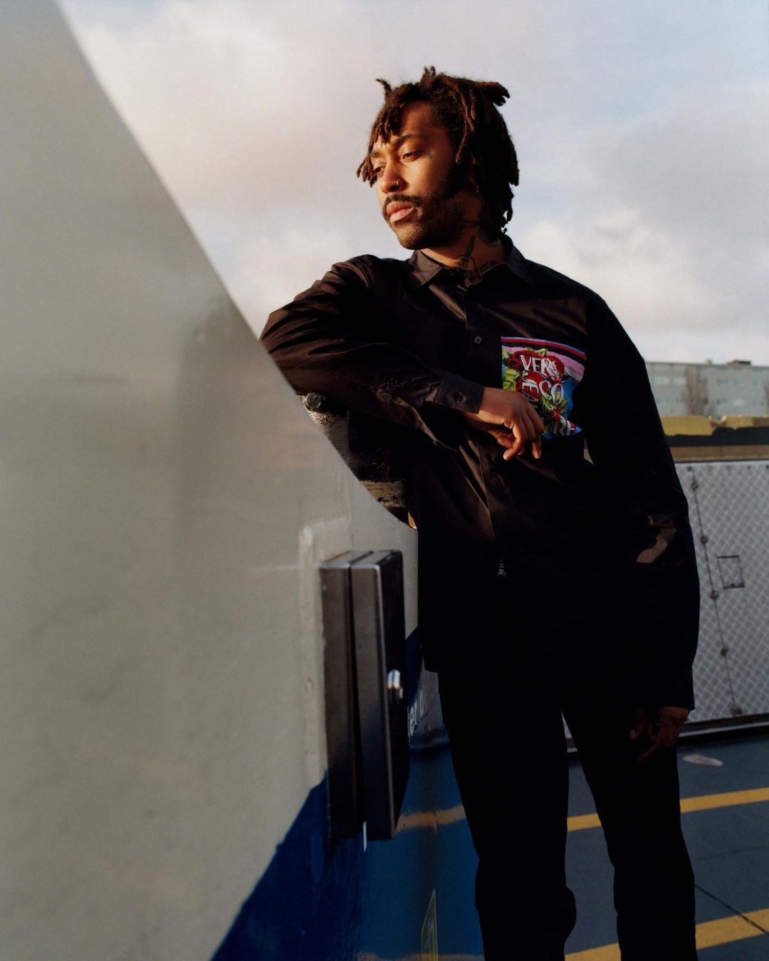 Adult male with dreadlocks leans against a metal structure outdoors on a cloudy day, wearing a dark jacket and pants.