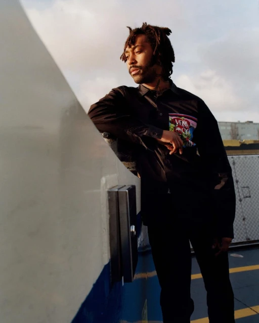 Adult male with dreadlocks leans against a metal structure outdoors on a cloudy day, wearing a dark jacket and pants.