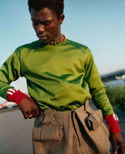 An adult Black male model poses outdoors wearing a shiny green long-sleeved shirt, tweed pants, and a gold chain necklace in natural light.