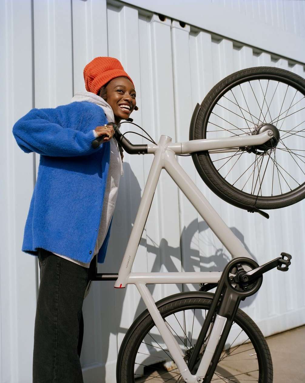 Young woman with a wide smile holds a light gray bicycle aloft against a corrugated metal wall, wearing a blue sweater and orange beanie.