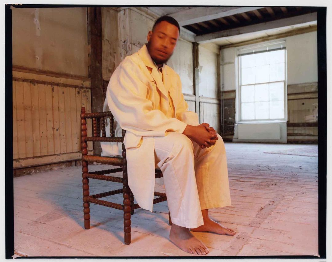 Portrait of a barefoot adult male in a cream suit sitting in an empty room with weathered walls and wooden floors.