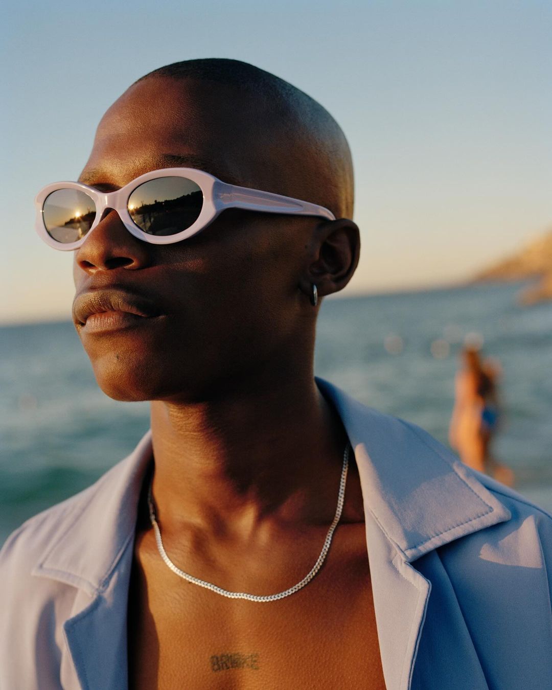 Close-up of a Black adult man wearing sunglasses and a silver chain, with a beach and other people visible in the background.