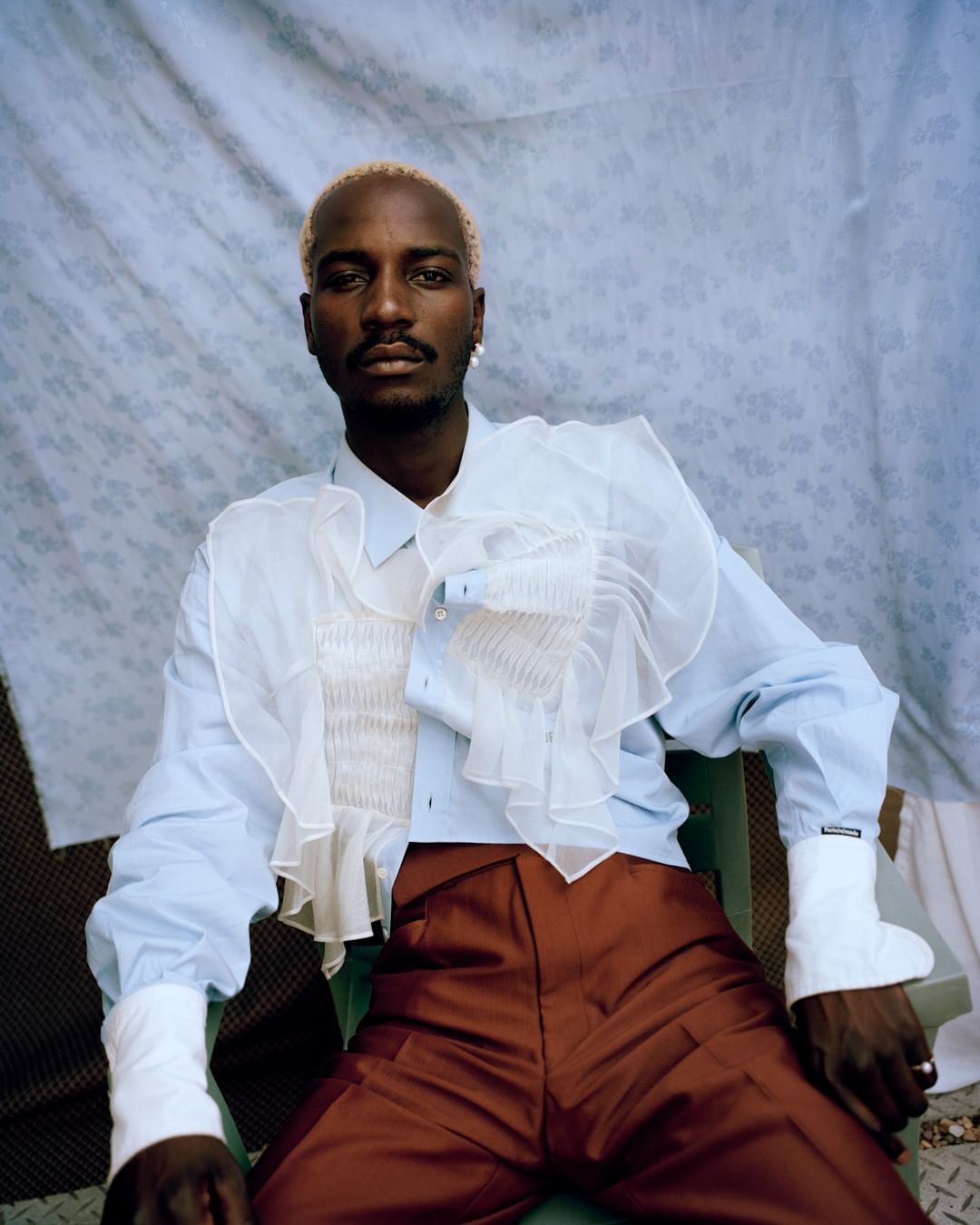 A Black male model with blond hair is photographed in a light blue ruffled shirt and red pants against a light blue patterned backdrop.