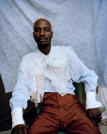 A Black male model with blond hair is photographed in a light blue ruffled shirt and red pants against a light blue patterned backdrop.