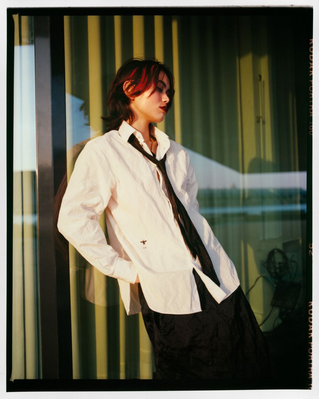 Woman with red-streaked dark hair in a white shirt and black tie poses against a window with vertical lines in golden light.