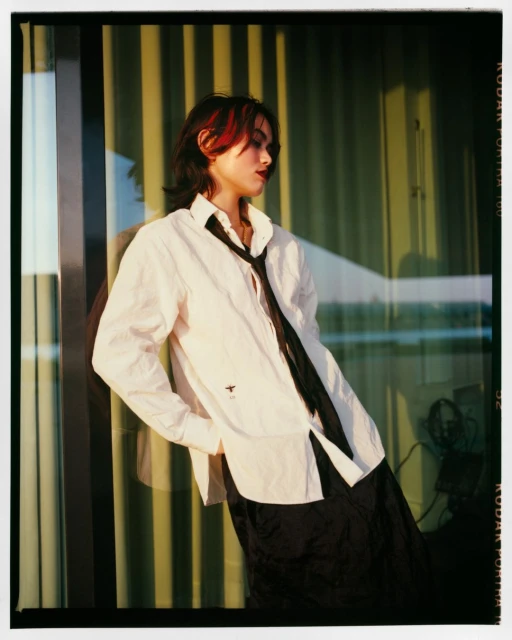Woman with red-streaked dark hair in a white shirt and black tie poses against a window with vertical lines in golden light.
