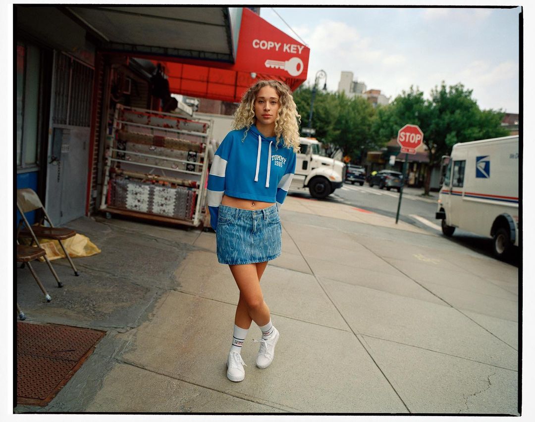 A young woman with curly blonde hair poses in a denim skirt and cropped sweatshirt on a city street with a postal truck.