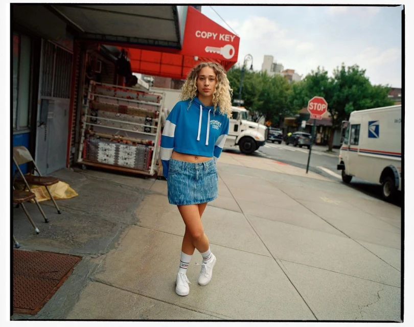 A young woman with curly blonde hair poses in a denim skirt and cropped sweatshirt on a city street with a postal truck.