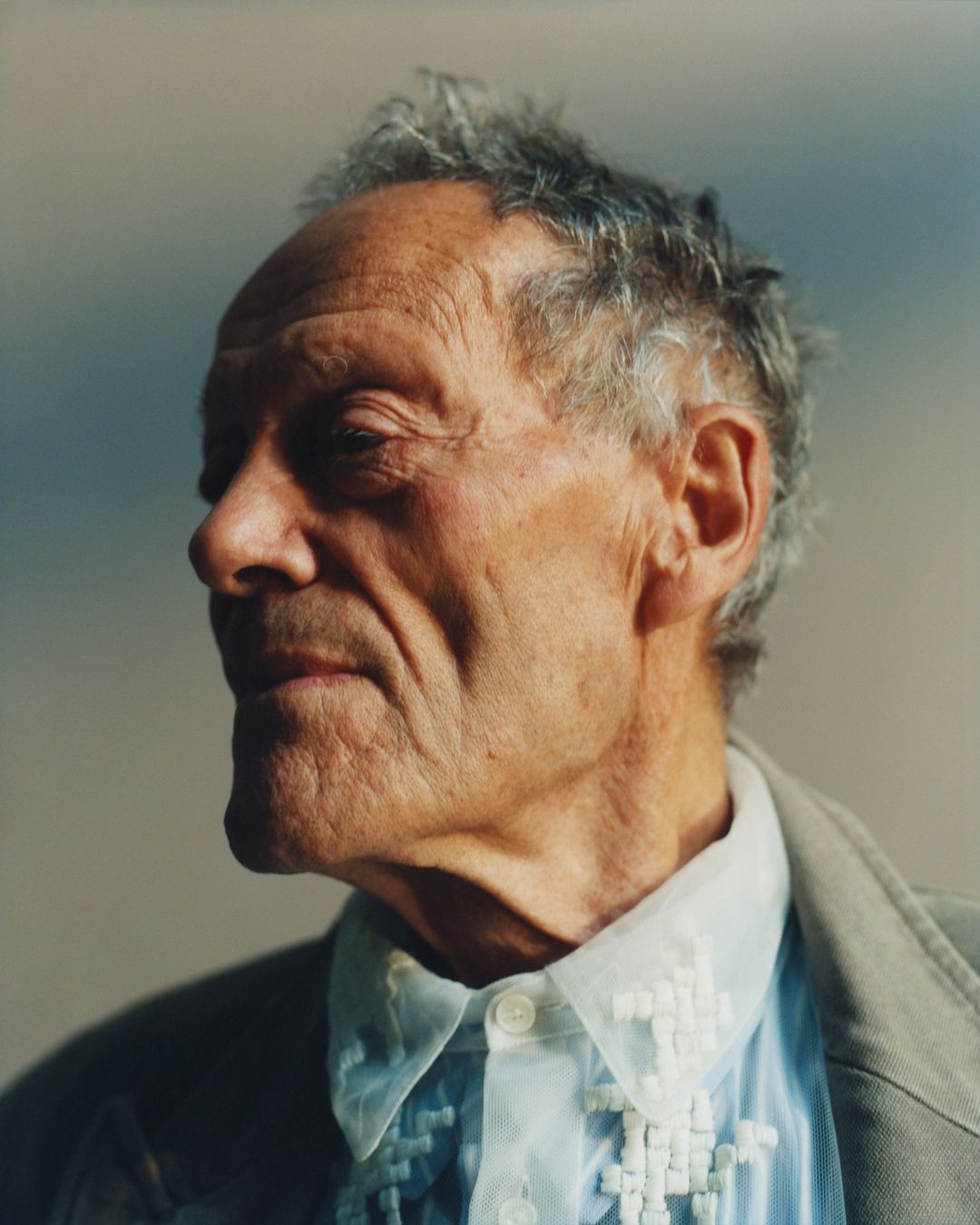 Portrait of an elderly man with gray hair wearing a patterned shirt and tan jacket against a neutral background in a medium shot.