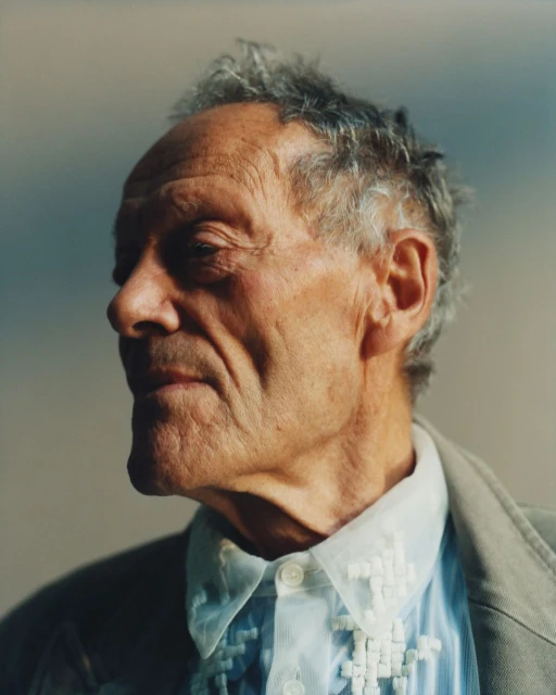 Portrait of an elderly man with gray hair wearing a patterned shirt and tan jacket against a neutral background in a medium shot.