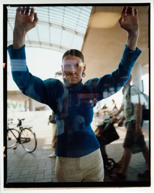 Woman with hands up stands behind glass pane with reflections of pedestrians and bicycle; modern architecture background.
