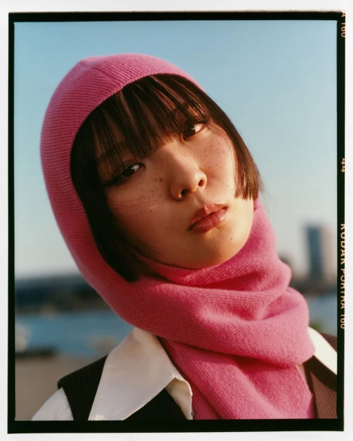 Portrait of a young woman with pink headscarf and neutral expression against a bright sky and blurred city background. Fashion.