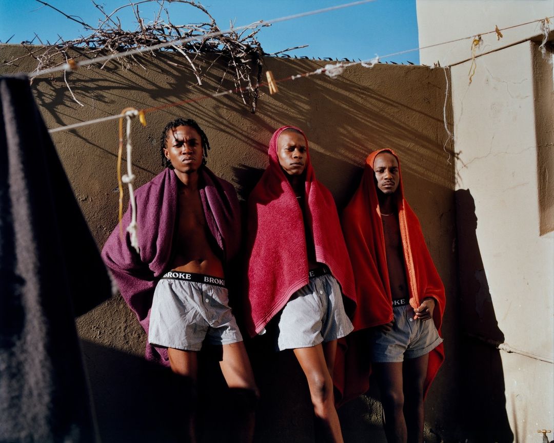 Three Black men pose draped in red and purple towels, wearing branded boxer shorts in an outdoor setting against a textured wall.