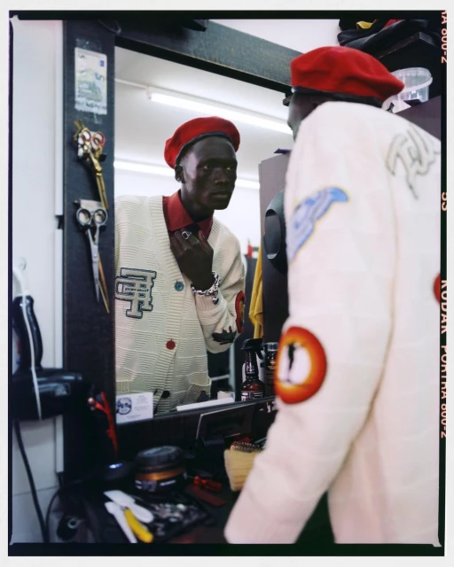 Man wearing a red beret, light-colored cardigan with patches, and jewelry, looking in the mirror of a barber shop.