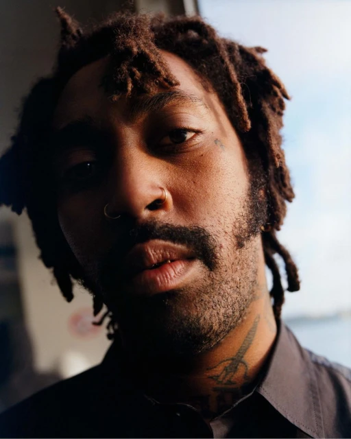 Portrait of a Black man with dreadlocks, a nose ring, and tattoos in a close-up shot, wearing a buttoned shirt, against a soft window light.