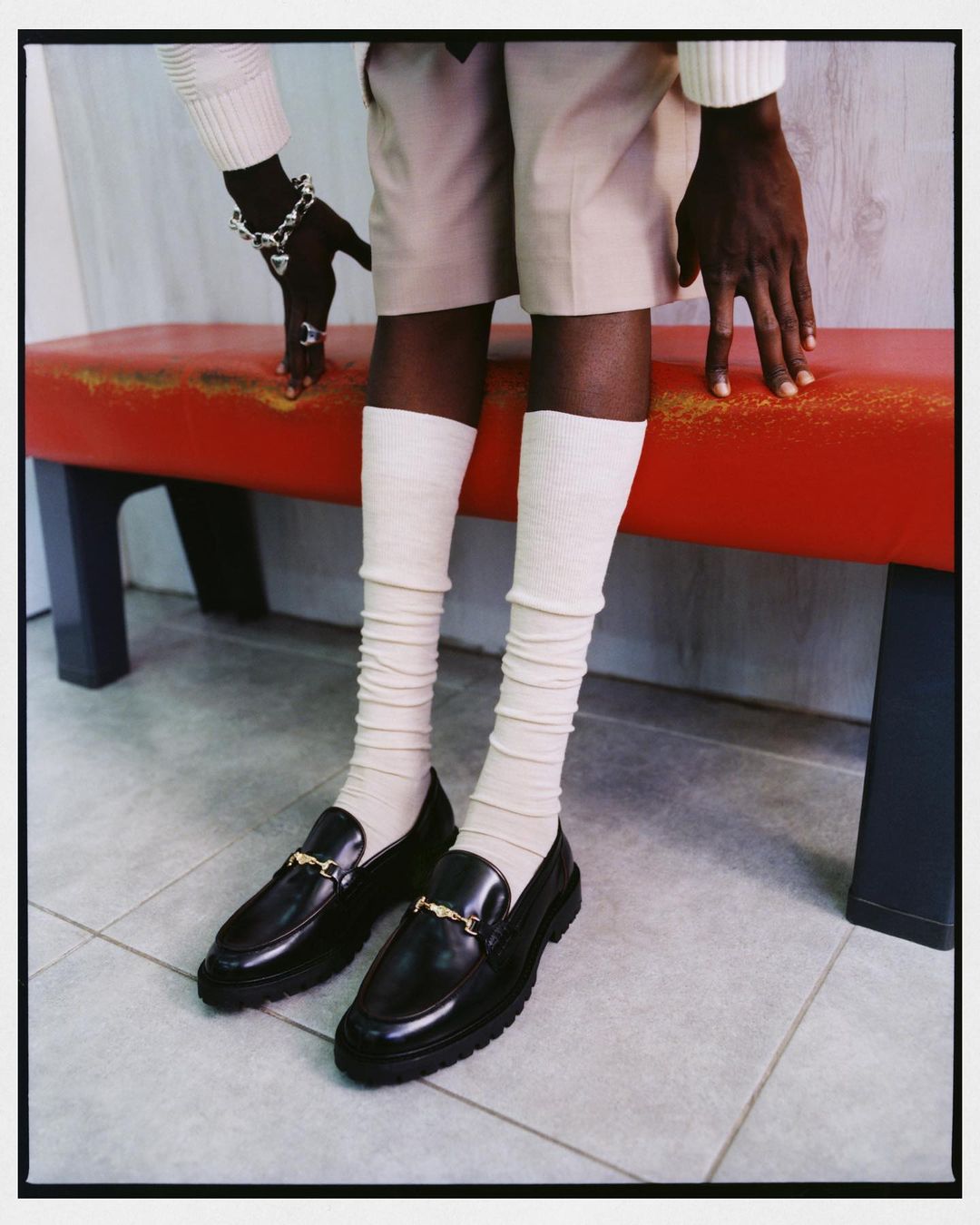 A person wearing black loafers with gold hardware, cream-colored socks and shorts, and a matching sweater sits on a red bench indoors.