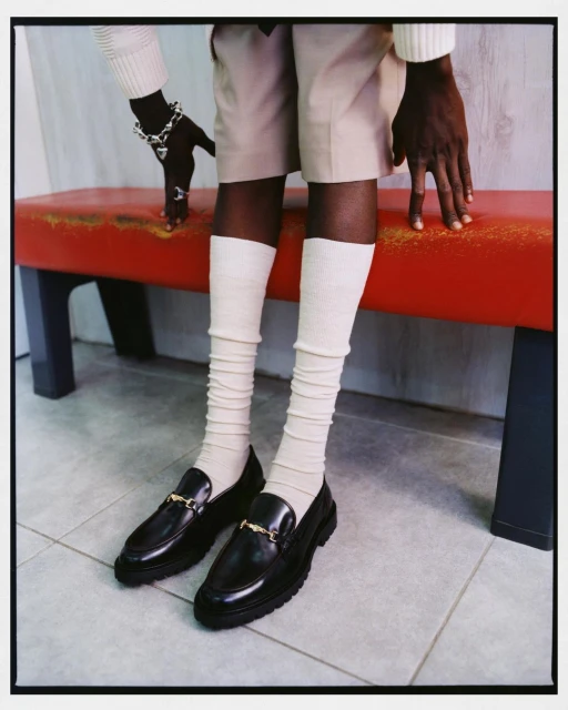 A person wearing black loafers with gold hardware, cream-colored socks and shorts, and a matching sweater sits on a red bench indoors.