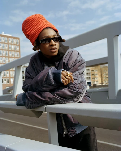 Stylish woman with orange beanie and sunglasses leans on railing with city buildings in background on sunny day. Fashion editorial shot.