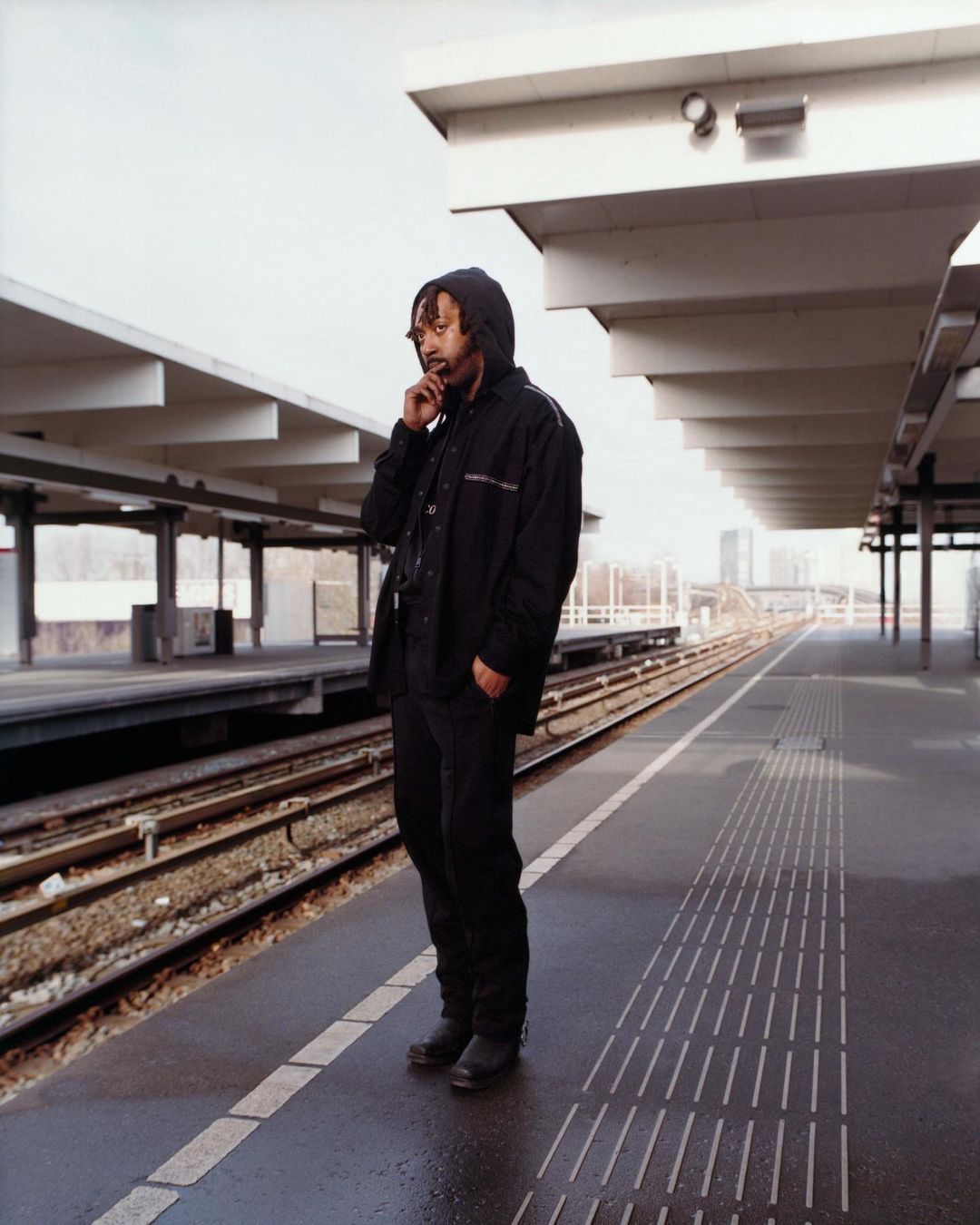 Full shot of an adult male in a hooded jacket standing on a train platform while using his smartphone on a cloudy day in an urban area.