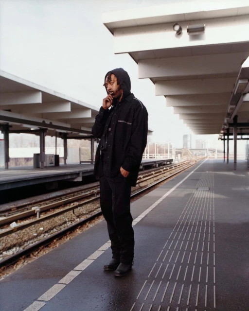 Full shot of an adult male in a hooded jacket standing on a train platform while using his smartphone on a cloudy day in an urban area.
