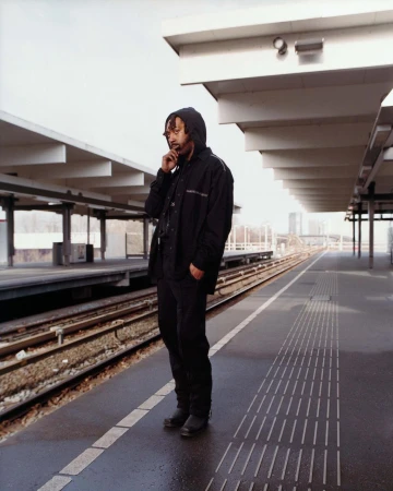 Full shot of an adult male in a hooded jacket standing on a train platform while using his smartphone on a cloudy day in an urban area.