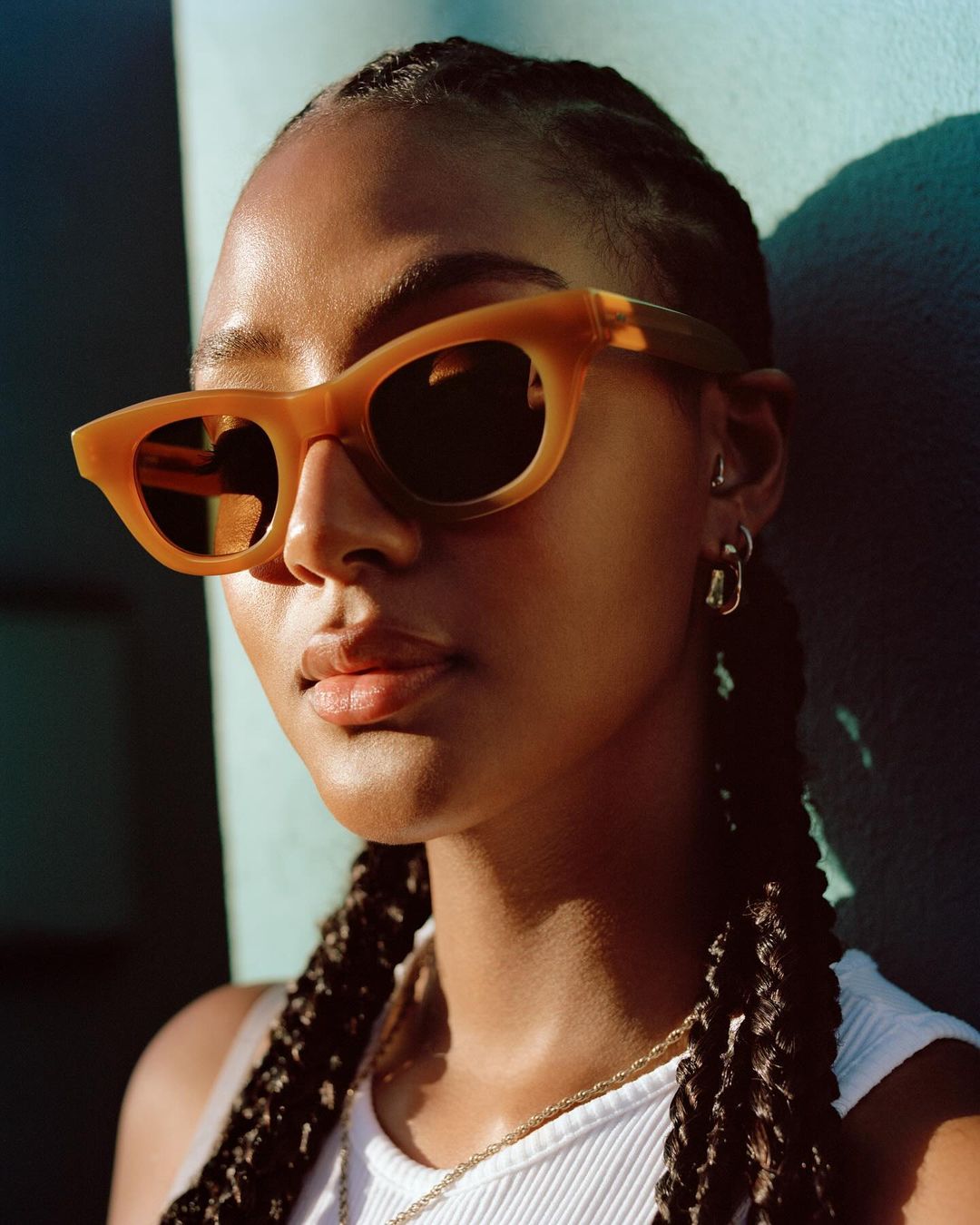 Close-up studio shot of a young adult female model wearing brown sunglasses with braided hair and a white top against a teal wall.