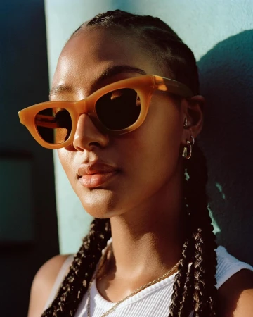 Close-up portrait of a woman wearing amber cat-eye sunglasses and a white ribbed tank top in direct sunlight.