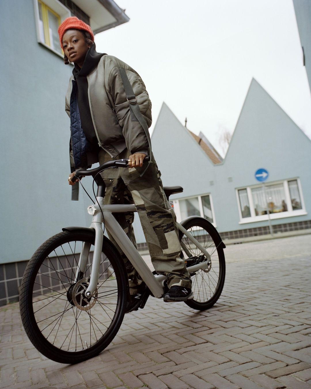 A young adult woman with a red beanie stands next to a modern gray bicycle on a brick street with residential buildings in the background.