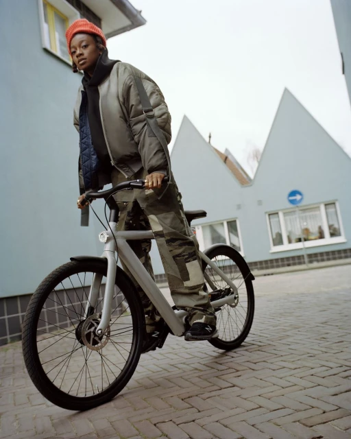 A young adult woman with a red beanie stands next to a modern gray bicycle on a brick street with residential buildings in the background.