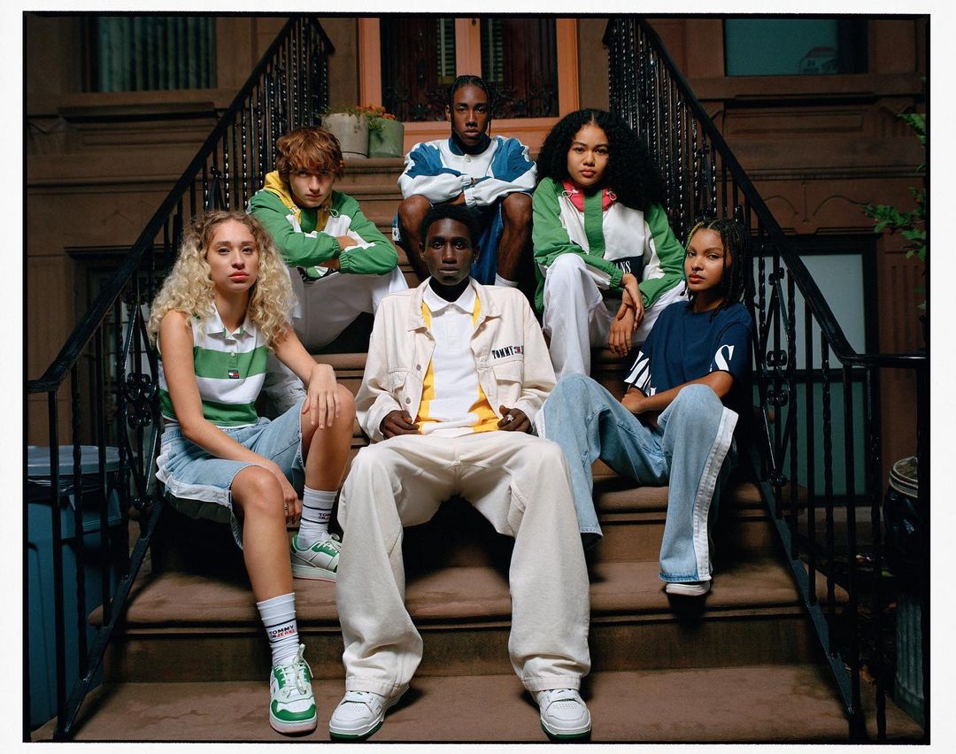 Group of six young adults posing on brownstone steps in 90's fashion apparel, showcasing a mix of casual and athletic styles with urban setting.