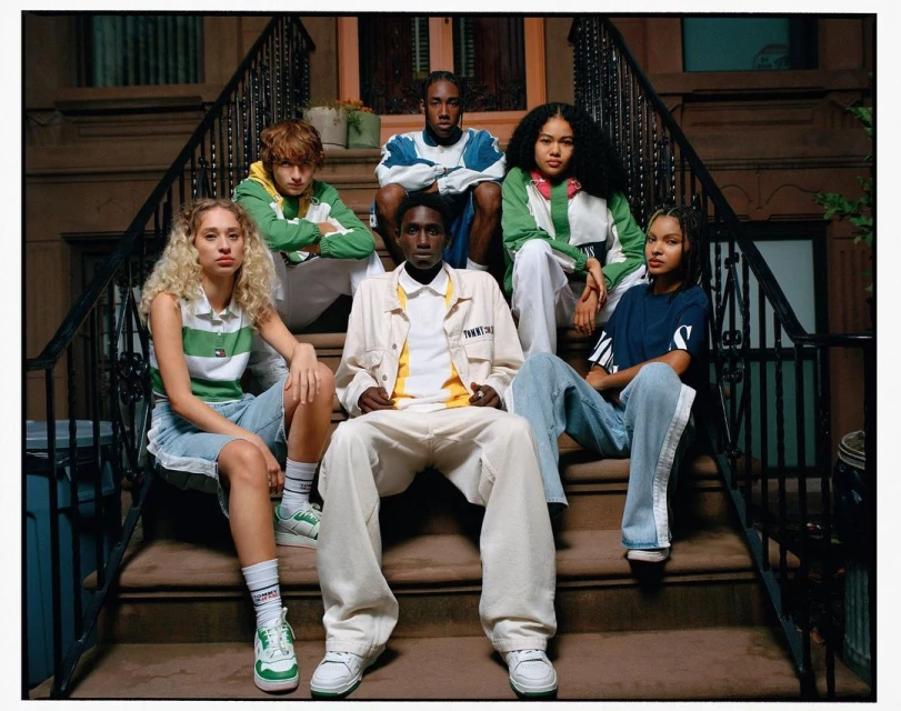 Group of six young adults posing on brownstone steps in 90's fashion apparel, showcasing a mix of casual and athletic styles with urban setting.