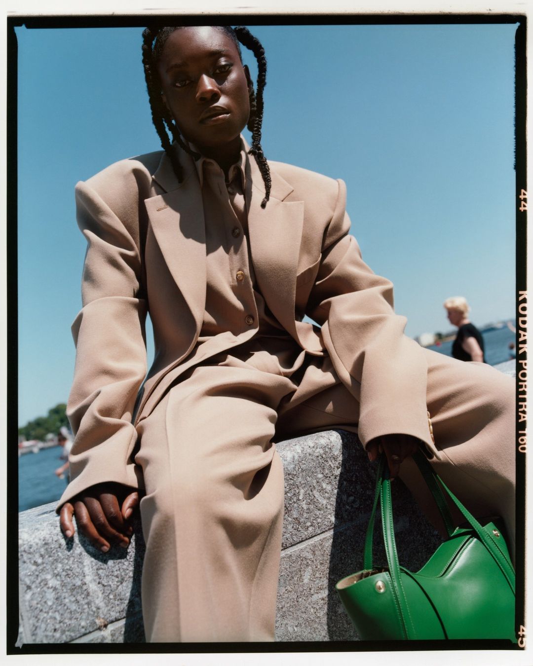 Fashion shot of a Black woman in a tan suit sitting on a stone wall near water, holding a green purse, with a blue sky backdrop.