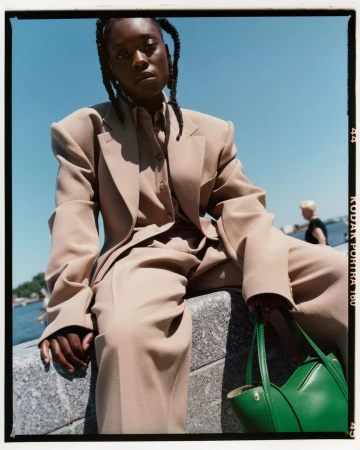 Black woman in beige tailored suit holding a green handbag sitting outdoors by water