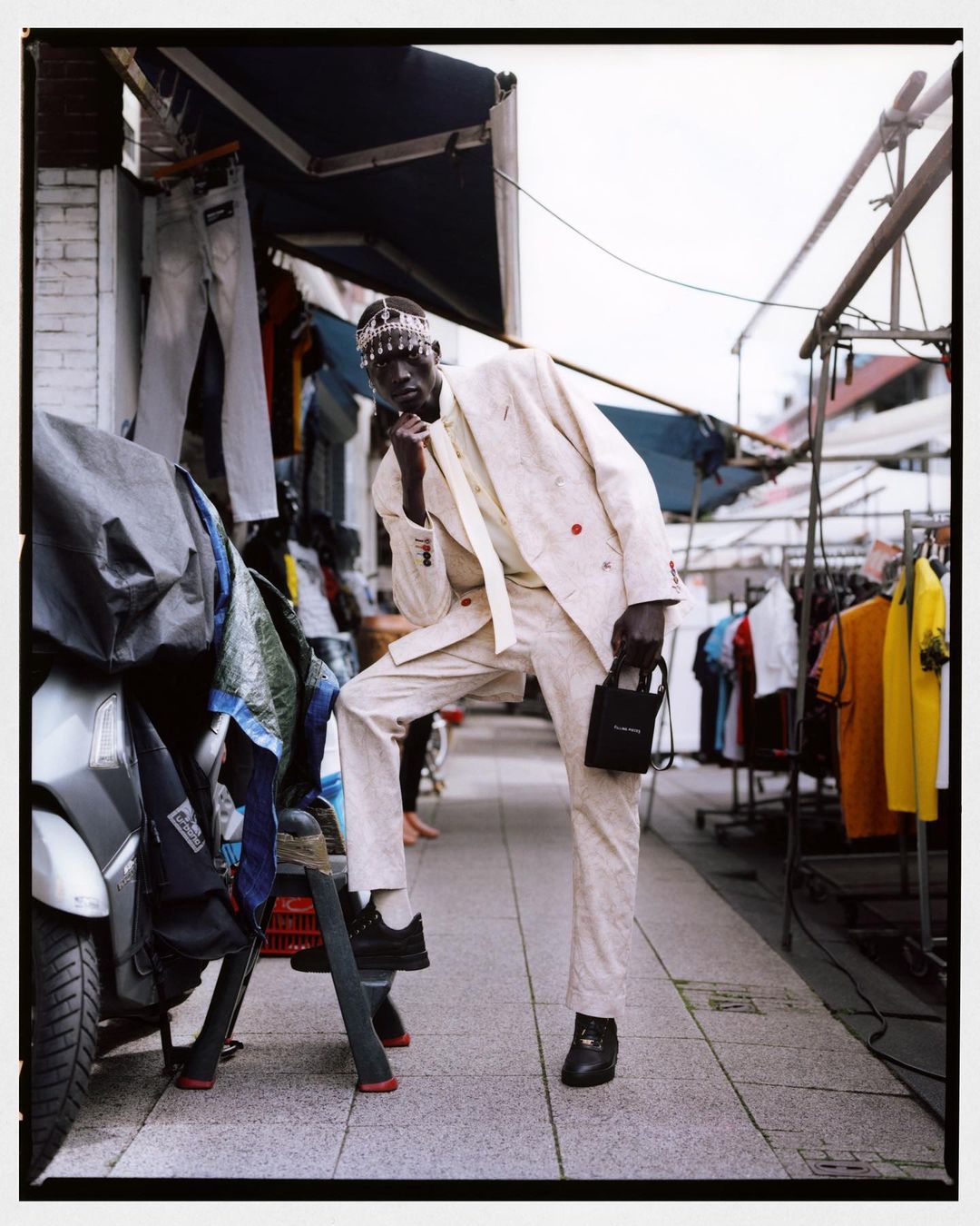 A stylish black man in a cream suit poses outdoors in an urban market with shirts and a moped, holding a small black handbag.