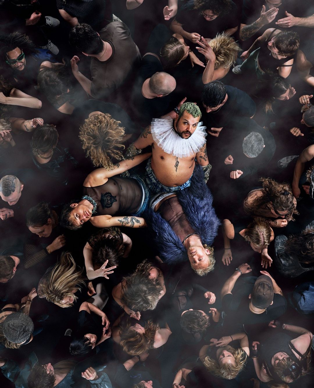 Overhead shot of a crowd with three people in the foreground: one in a ruffled collar, and two others lying down.