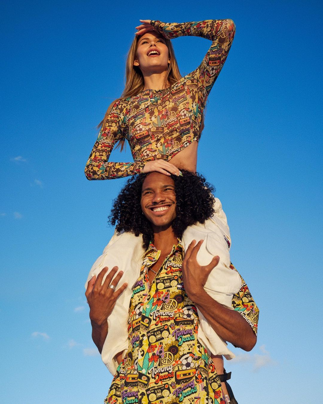 Young adult couple enjoying a sunny day, woman with long hair sits on man's shoulders against a clear blue sky. Both are smiling.