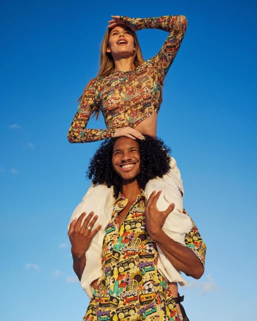 Young adult couple enjoying a sunny day, woman with long hair sits on man's shoulders against a clear blue sky. Both are smiling.