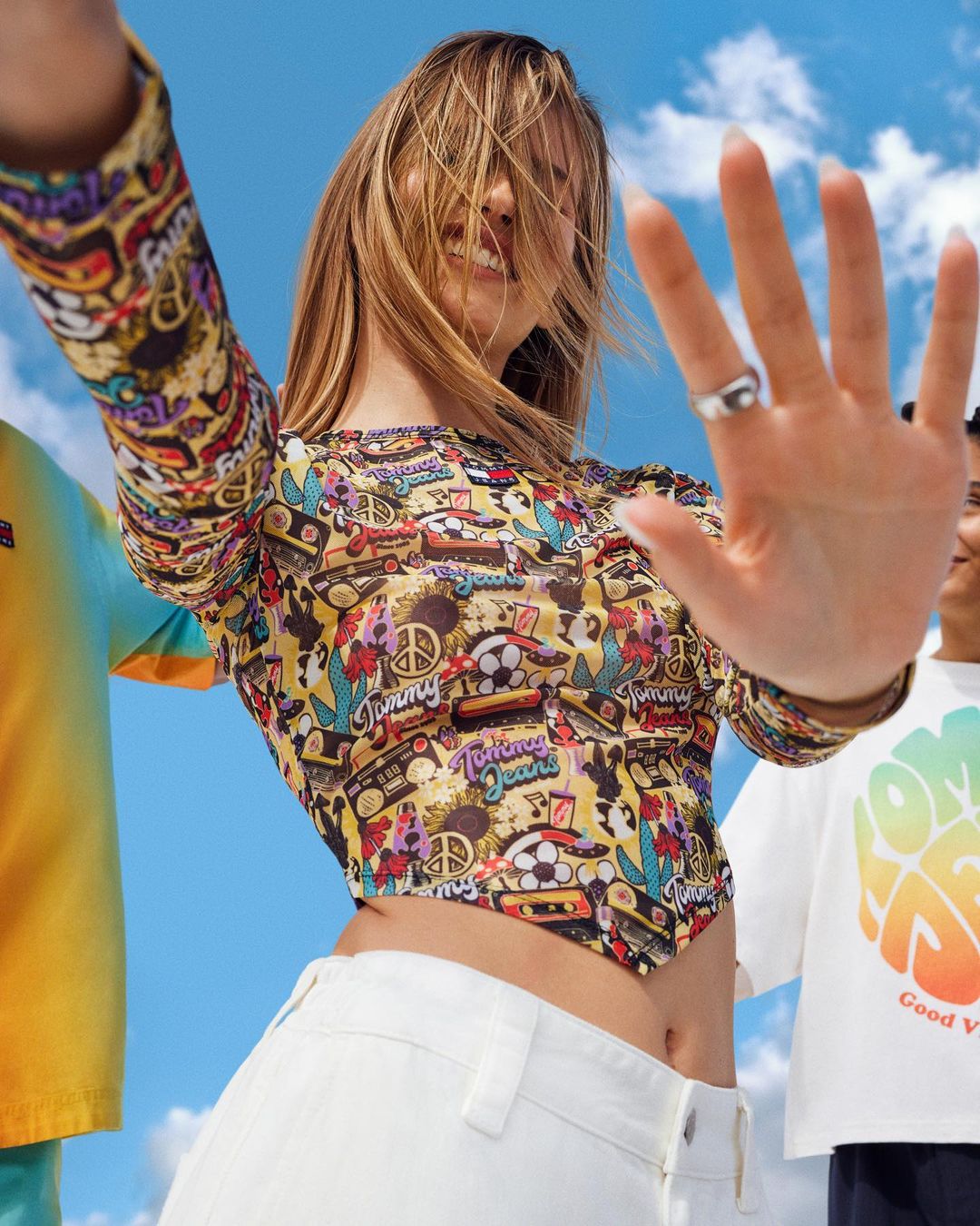 A young woman smiles in a patterned top and white pants, holding up a hand, with people and blue sky visible in the background.