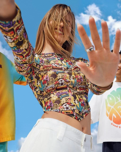 A young woman smiles in a patterned top and white pants, holding up a hand, with people and blue sky visible in the background.