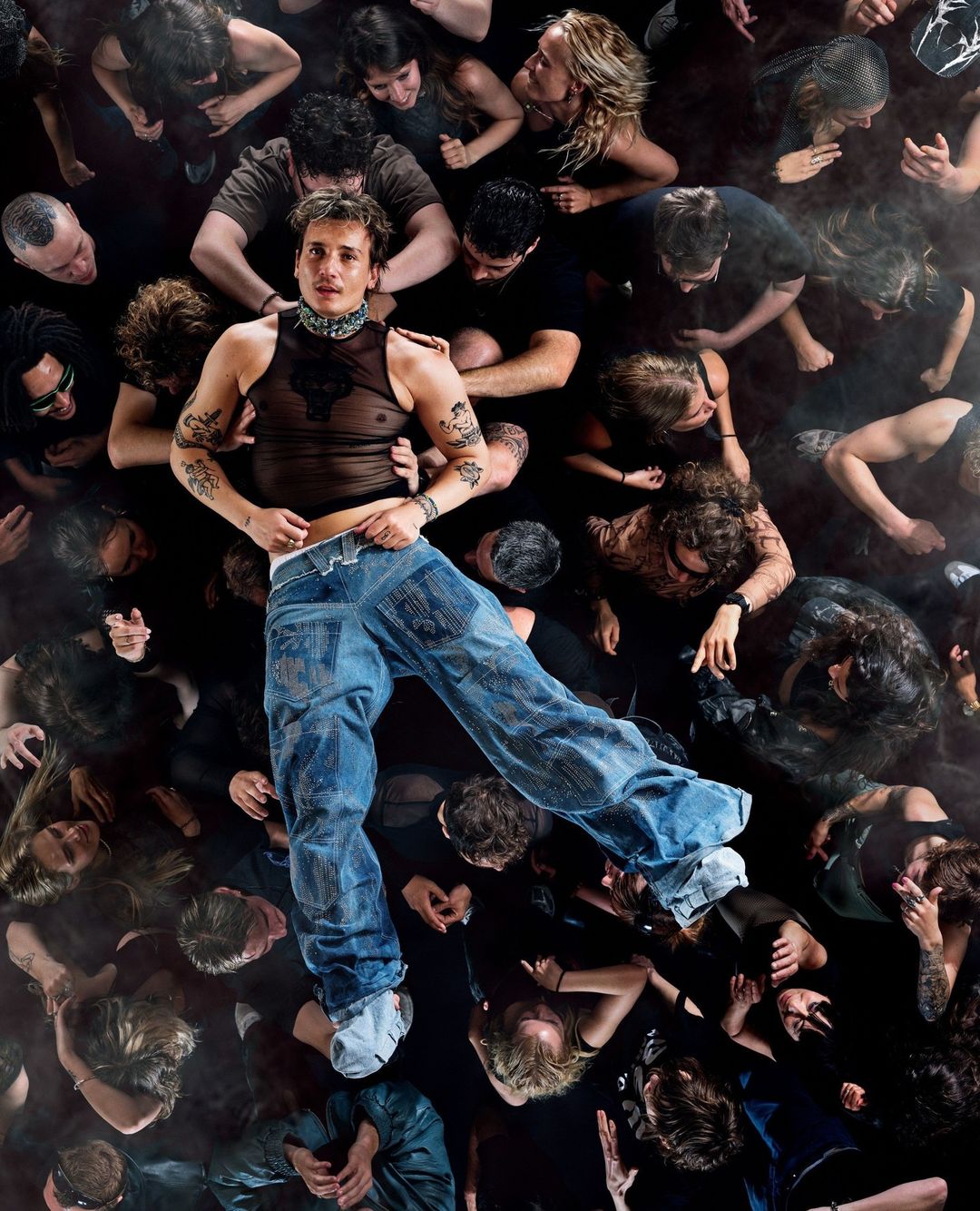 Aerial view of a fair-skinned adult male wearing a sheer top and blue jeans standing out from a crowd of diverse adults looking up.