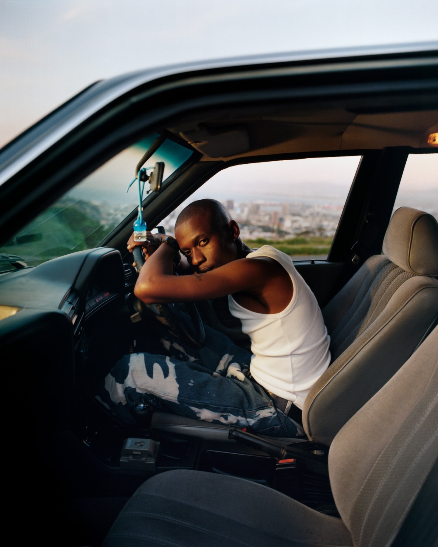 A young man with a shaved head leans against the steering wheel in the driver's seat of a car against a distant cityscape backdrop.