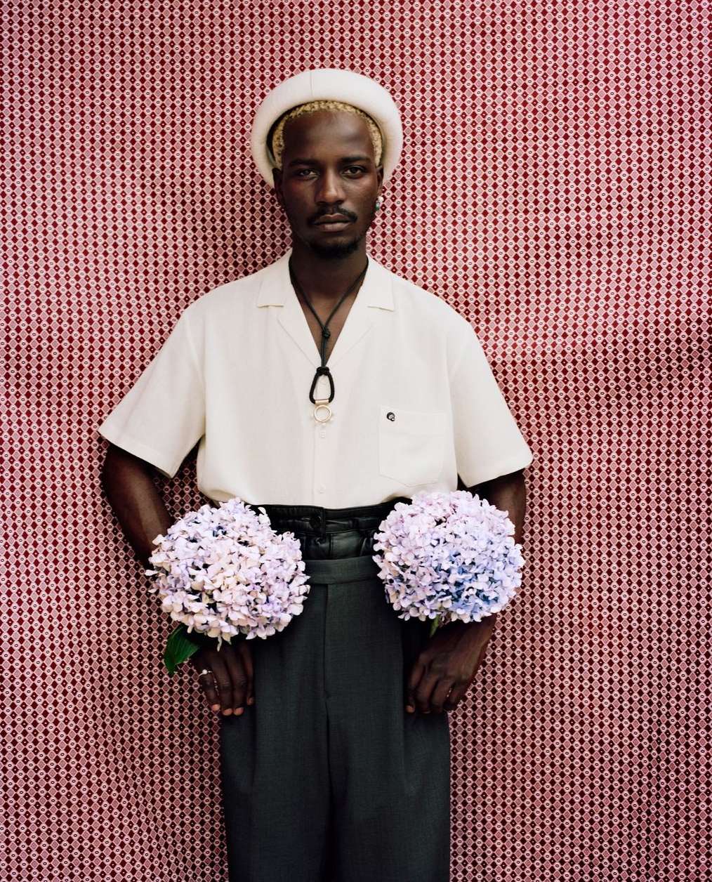 Adult male with short blond hair wearing a cream-colored hat, cream shirt, and black pants, holding hydrangeas in front of red patterned background.