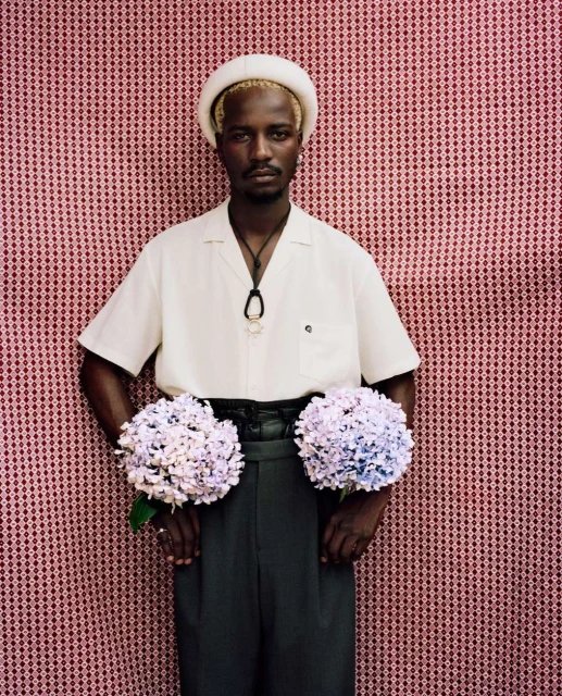 Adult male with short blond hair wearing a cream-colored hat, cream shirt, and black pants, holding hydrangeas in front of red patterned background.