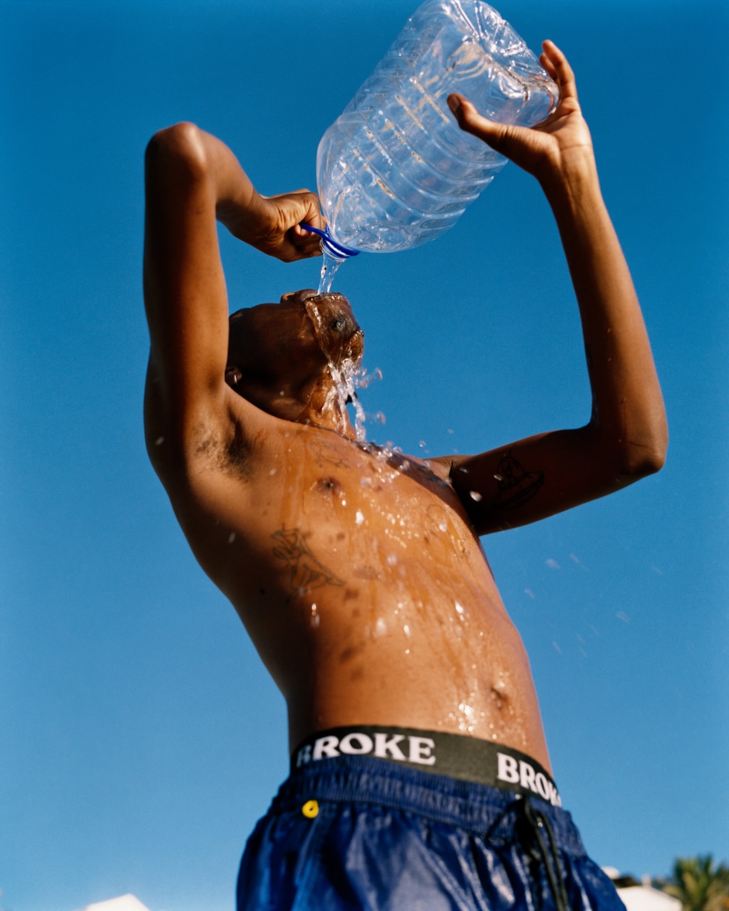 Adult male pouring water from a transparent plastic bottle over his head in bright sunlight with a clear blue sky in the background.